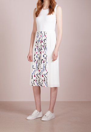 Witte mouwloze jurk met een geplooide rok met een kleurrijke abstracte print. Gecombineerd met witte sneakers, tegen een neutrale achtergrond.