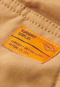 Etiqueta Superdry Surplus sobre tela marrón. La etiqueta rectangular presenta un fondo naranja con texto negro. La textura es tejida y suave.