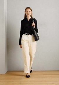 Camicia nera con bottoni, pantaloni bianchi a gamba larga, scarpe basse nere e una borsa tote scura, su uno sfondo neutro con pavimento in legno.