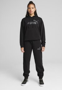 Juodas džemperis su kišene priekyje ir PUMA logotipu ant krūtinės, derinamas su juodomis treniruotėmis, turinčiomis logotipo detalę, ir juodais sportiniais batais.