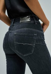 Primer plano de una persona que lleva puestos unos vaqueros de mezclilla azul oscuro con una etiqueta plateada de "Salsa Jeans" en la cintura y bolsillos traseros con costuras.