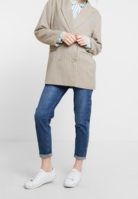 Blazer doppiopetto beige a righe, camicia a righe blu chiaro, jeans blu e sneakers bianche. Il blazer presenta grandi bottoni e una vestibilità rilassata.