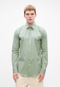 Um jovem com cabelo castanho claro curto usa uma camisa de botão verde-claro e calças creme, de pé contra um fundo branco simples.