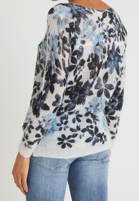 Pull en tricot à motif floral avec une texture douce, présentant des fleurs bleues et noires sur un fond crème, avec un large décolleté et un design à épaules tombantes.