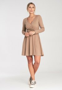 Beżowa sukienka z długim rękawem, o dopasowanej górze i rozszerzanym dole, w zestawieniu z białymi trampkami w czarne plamki.