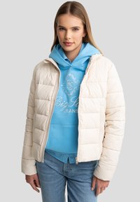 Elfenben pufferjakke med quiltet struktur, med frontlynlås og stående krave, båret over en lyseblå hættetrøje og denimjeans.
