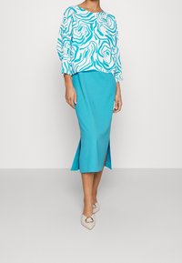 Blusa a maniche lunghe con stampa floreale blu abbinata a una gonna turchese a tinta unita con spacchi laterali. Scarpe beige a punta con dettagli in metallo.