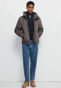 Graue Kapuzenjacke mit Frontreißverschluss, kombiniert mit einem marineblauen Strickpullover, blauen Jeans und beigen Schuhen. Einfaches Design, lässige Passform.