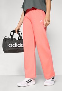 Pink sweatpants with a relaxed fit, paired with a dark gray top, white sneakers, and a black duffel bag featuring bold white branding.