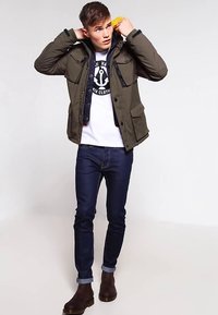 Grøn vatteret jakke med sorte detaljer, fire forlommer, båret over en hvid T-shirt. Mørke jeans og brune ankelstøvler fuldender outfittet.