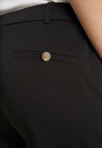 Pantaloni neri con una texture liscia, una leggera lucentezza e una tasca posteriore chiusa con un bottone metallico che mostra un logo.