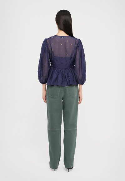Blusa semitranslúcida azul marino con mangas abullonadas, textura estampada, cintura ajustada, combinada con pantalones verdes de talle alto y tacones negros.