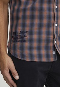 Camisa de cuadros de manga corta en tonos azules, naranjas y grises, con botones blancos y un bordado de la marca en el dobladillo. Corte estándar.