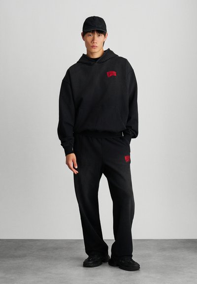 Conjunto de sudadera negra con capucha y pantalones de chándal, que presenta un pequeño logo rojo en el pecho y en los pantalones. Material de algodón con un corte relajado y capucha ajustable.