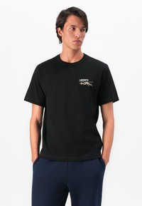 Un jeune homme portant un t-shirt noir Lacoste avec un petit graphique de montagne sur la poitrine, les mains dans les poches de son pantalon bleu marine, regardant à droite.