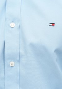 Chemise bleu clair à l'avant avec des boutons blancs et un petit logo rectangulaire présentant des rayures horizontales rouges, blanches et bleues.