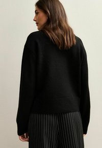 Maglione nero a maglia con una vestibilità comoda, caratterizzato da un collo rotondo e orlo a coste. Indossato con pantaloni a righe e capelli texturizzati in onde morbide.