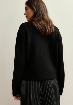 Jersey negro de punto con un corte relajado, que presenta un cuello redondo y un dobladillo acanalado. Combinado con pantalones a rayas y el cabello texturizado en ondas sueltas.