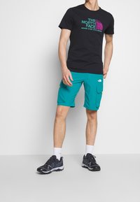 Svart t-shirt med kort ärm och färggård logotyp, tillsammans med turkosa shorts med fickor och ett andningsbart tyg. Svarta sneakers.