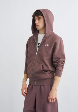 Mladý muž v mauve mikine so zipsom a logom Nike, s nohavicami v rovnakej farbe a bielom tričku Nike, stojaci na jednoduchom svetlom pozadí.