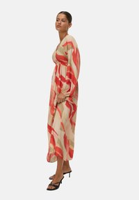 Femme portant une longue robe beige avec des motifs abstraits rouges et roses et des sandales à talons hauts noires à lanières, se tenant de côté.