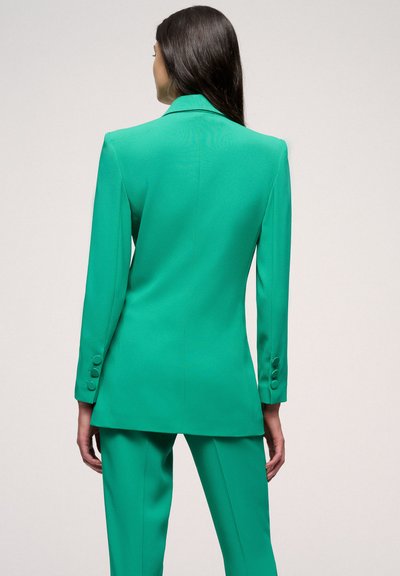 Groene blazer en broek set van soepel materiaal. De blazer heeft een scherpe kraag en drieknoopmanchetten, met een op maat gemaakte pasvorm.