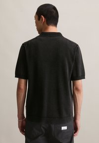 Mężczyzna widziany z tyłu, ubrany w czarną, krótkosleeve'ową dzianinową koszulę polo i czarne spodnie z białą metką na tylnej kieszeni.