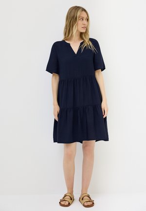 Marineblaues Baumwollkleid mit kurzen Ärmeln, einem V-Ausschnitt und drei Lagen, das einen entspannten Schnitt und eine weiche Textur aufweist, kombiniert mit braunen Sandalen.