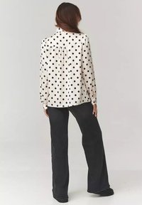 Bluzka z długim rękawem w kolorze kremowym z dużymi czarnymi kropkami, z tylnym karczkiem i luźnym krojem, w połączeniu z czarnymi spodniami o szerokich nogawkach.