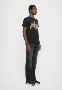 Črna majica s kratkimi rokavi in pisanimi napisi "VERSACE JEANS COUTURE". Sive denim hlače z rahlim razširjenjem. Črne športne copate z belimi detajli.