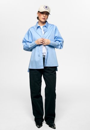 Person som har på seg en beige caps, en løs, lyseblå skjorte med knapper over en hvit topp, svarte bukser og svarte spisse sko, stående mot hvit bakgrunn.