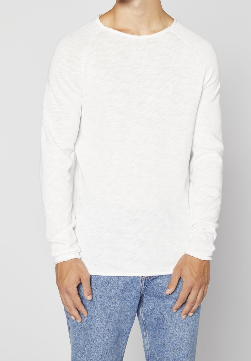 Vit långärmad skjorta i ett lätt, strukturerat tyg med rundad halsringning och raglanärmar, parat med blå denimjeans.