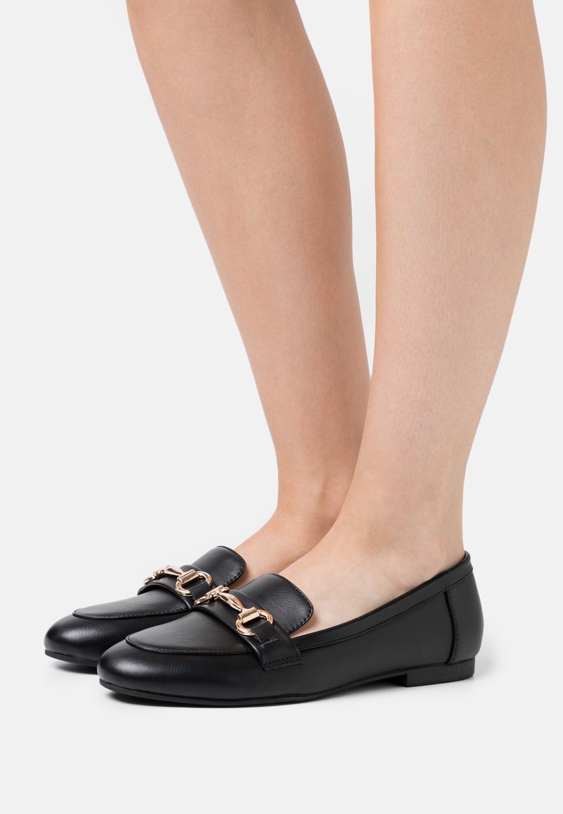 Des mocassins en cuir noir présentent un design épuré, des accents en chaîne dorée à l'avant et une semelle plate. Le matériau a une texture lisse.