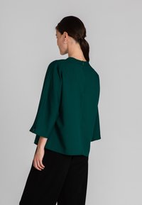 Blusa verde, strutturata, con un collo alto, maniche a tre quarti e un dettaglio con cerniera sul retro, abbinata a pantaloni neri.
