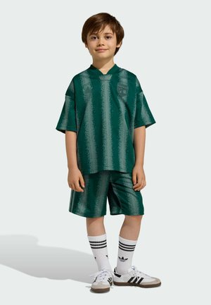 Junge, der steht und ein grünes, gestreiftes Sporttrikot und Shorts trägt, dazu weiße Adidas-Socken und Sneakers, vor hellem Hintergrund.