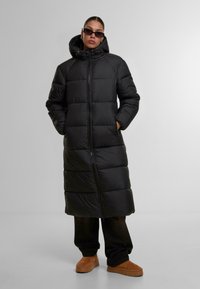 Svart puffjacka med en lång, vadderad design, dragkedjestängning, huva och sidofickor. Bärs över mörka byxor och bruna slip-on-skor.