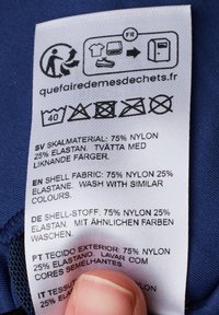 Tvättanvisning på ett blått tyg. Innehåller fiberkomposition: 75% nylon, 25% elastan, tvättinstruktioner och återvinningsikoner.