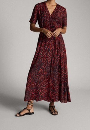 Woman wearing a black and red patterned wrap dress with short sleeves and black strappy flat sandals, standing on a gray background.