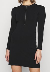 Femme portant une robe noire à manches longues avec une fermeture éclair argentée à l'avant et des poignets côtelés, montrée de la nuque jusqu'au milieu de la cuisse.