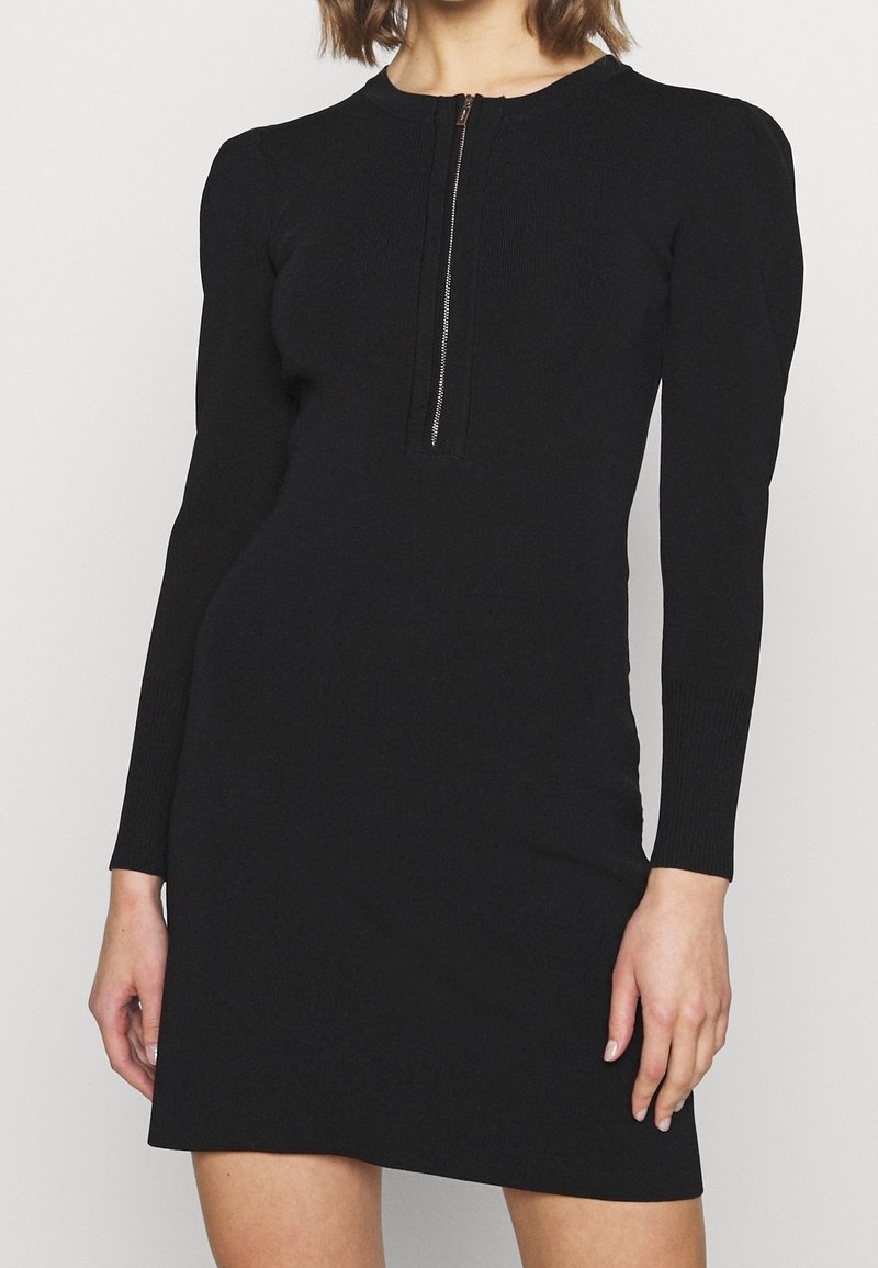 Femme portant une robe noire à manches longues avec une fermeture éclair argentée à l'avant et des poignets côtelés, montrée de la nuque jusqu'au milieu de la cuisse.