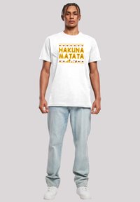 Hvit bomulls t-skjorte med "HAKUNA MATATA" i gul fet skrift, med dyregrafikk og stamme mønstre. Kombinert med lyseblå jeans og hvite sneakers.