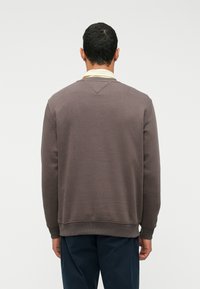 Uomo visto di spalle che indossa un felpa marrone sopra una camicia color giallo chiaro con colletto e pantaloni blu scuro, su uno sfondo bianco.