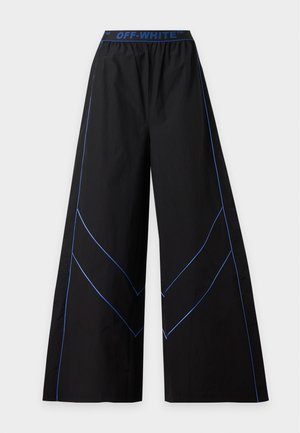 Femme debout, bras croisés, vêtue d’un pantalon noir à jambes larges et d’un top court sans manches avec un liseré bleu et la marque Off-White.