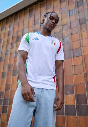 Biała koszulka piłkarska z krótkim rękawem z niebieskimi, czerwonymi i zielonymi akcentami, z logo flagi Włoch i teksturowanym wzorem. Noszona z jasnymi spodniami dżinsowymi.