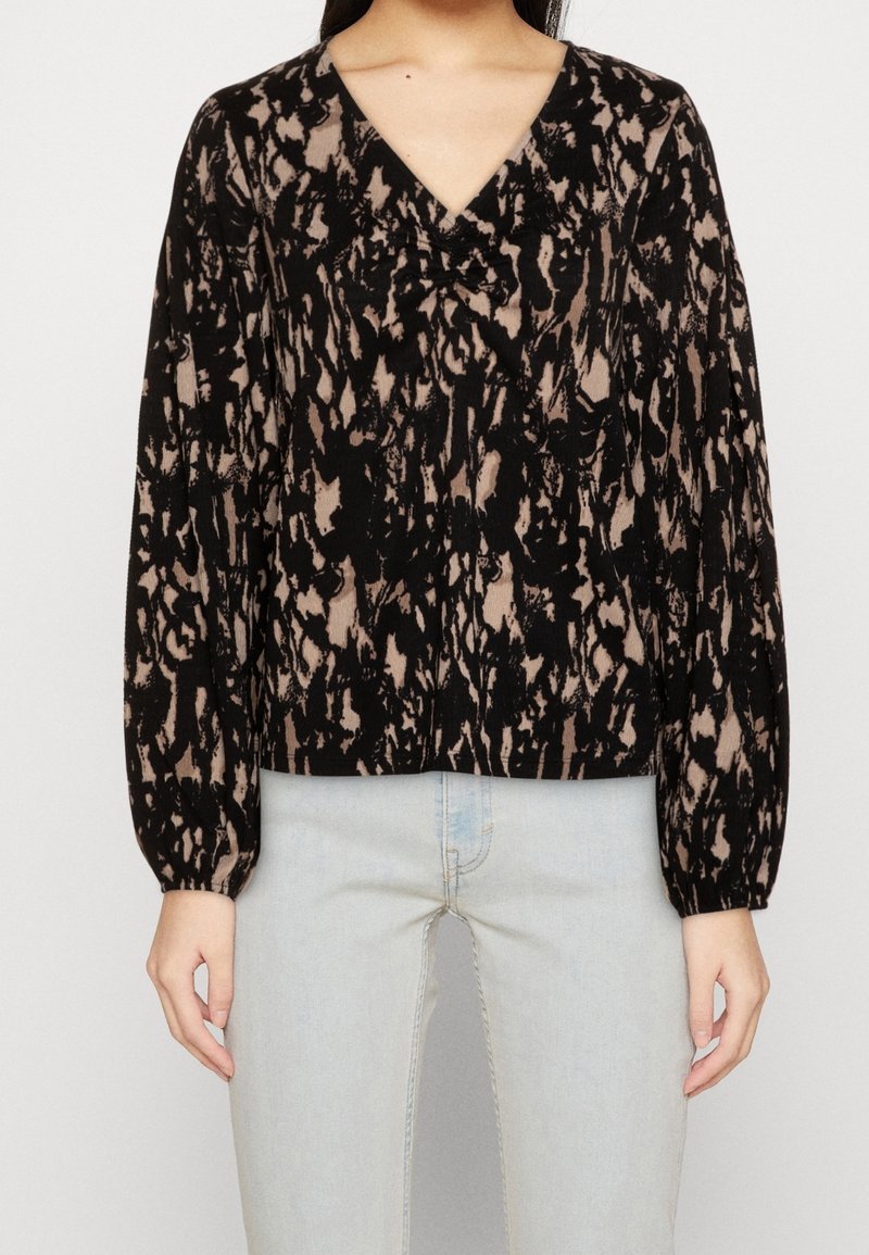 Schwarz-beige gemusterte Bluse mit V-Ausschnitt, gerafftem Vorderteil und langen Puffärmeln, kombiniert mit hellblauen Jeans. Weiche Textur.