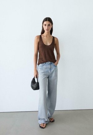 Femme debout contre un mur blanc, portant un haut marron sans manches, un jean large bleu clair, des sandales noires et tenant un petit sac à main noir.