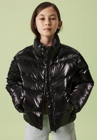 Giacca imbottita nera con finitura lucida e trapuntata, collo alto e cerniera frontale. Indossata sopra una maglia marrone e abbinata a jeans chiari.