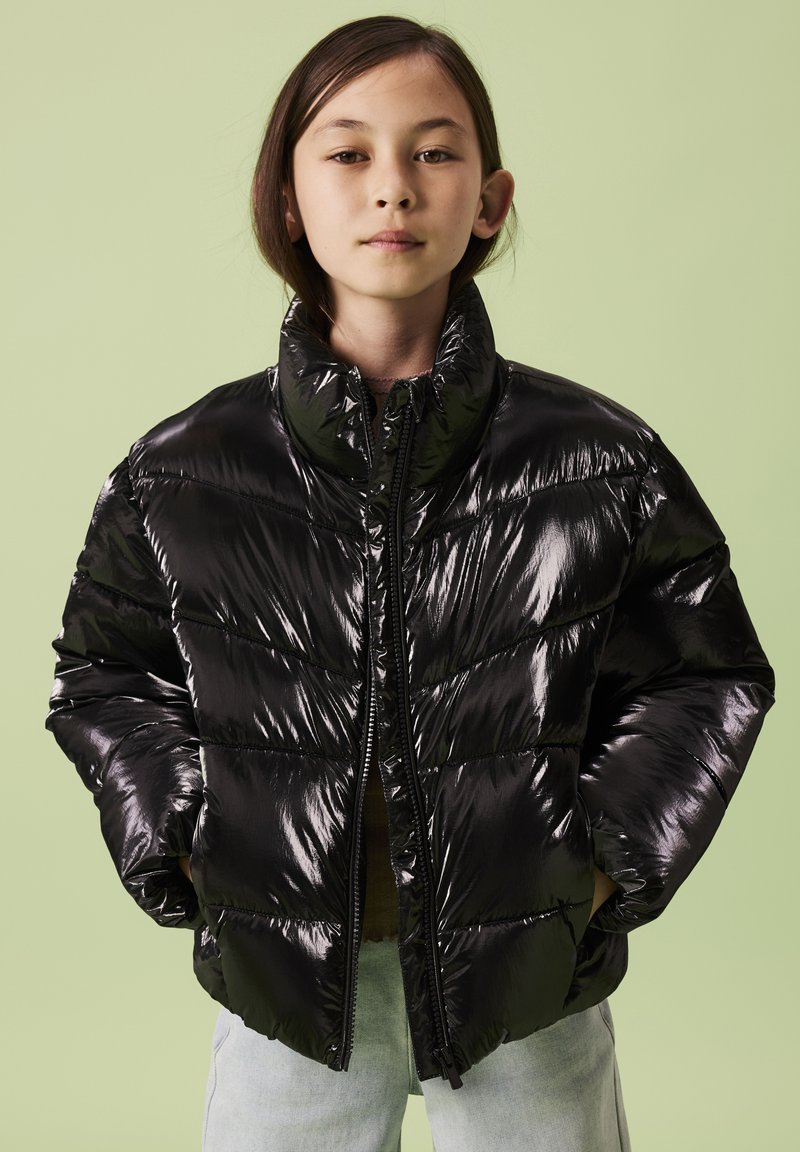 Giacca imbottita nera con finitura lucida e trapuntata, collo alto e cerniera frontale. Indossata sopra una maglia marrone e abbinata a jeans chiari.