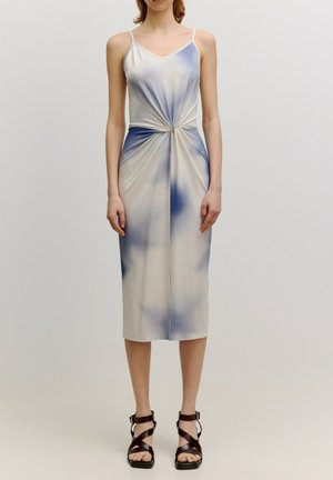 Woman wearing a sleeveless, light-colored midi dress with blue tie-dye pattern and twisted knot detail at the waist, paired with dark strappy sandals.