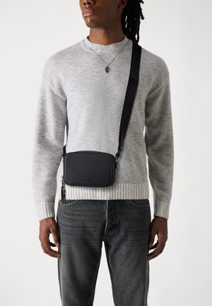 Borsa a tracolla in pelle nera con forma rettangolare, chiusura con zip e tracolla regolabile, indossata su un maglione grigio a coste e jeans scuri.